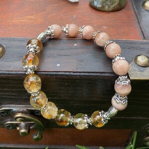 May include: A beaded bracelet featuring alternating peach and amber-colored gemstones, separated by ornate silver-toned metal beads. The bracelet is displayed on a wooden surface with other crystals and a decorative ceramic piece in the background.