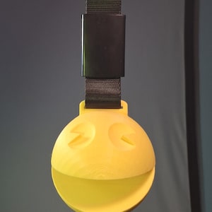 May include: A yellow, spherical object with a Pac-Man-like design, featuring two eye-shaped cutouts and a wide, open mouth. It is suspended by a black strap and a rectangular black clasp. The background is a gradient of gray and blue.