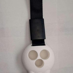 Puede incluir: Un llavero blanco con forma de calavera con tres cuencas oculares circulares y tres dientes puntiagudos. Está unido a una correa negra y un cierre rectangular negro. El llavero es probablemente de plástico.