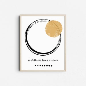 May include: A framed art print featuring a black circle and a gold-colored circle on a white background. The text "in stillness lives wisdom" is below the circles, with a row of black dots beneath the text. The frame is light-colored.