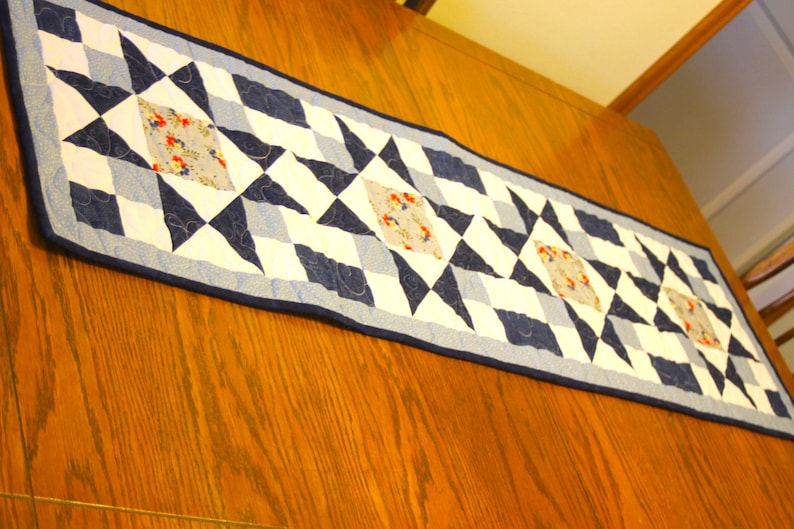 Puede incluir: Un camino de mesa acolchado azul y blanco con un patr&oacute;n geom&eacute;trico de estrellas. El camino de mesa tiene un borde azul claro y peque&ntilde;os cuadrados de tela floral en el centro de cada estrella.