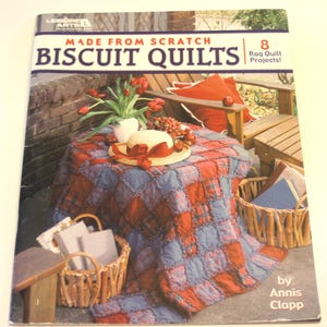 May include: A book cover titled "Made From Scratch Biscuit Quilts" with a red and blue quilt on a table. The scene includes a straw hat, flowers, and baskets, suggesting a quilting theme. The book promises 8 Rag Quilt Projects!