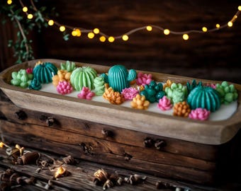 May include: A wooden trough candle filled with white wax and colorful succulent-shaped candles. The succulents are various shades of green, teal, orange, and pink. The candle sits on a wooden surface, with string lights in the background.