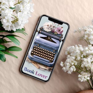 Pode incluir: Um smartphone exibe uma ilustração digital de uma máquina de escrever azul vintage com um arranjo floral. A tela mostra o texto "Book Lover". Flores brancas e roxas são dispostas na máquina de escrever.