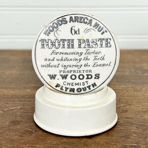 May include: A vintage white and black tooth paste container. The container has a circular top with the text "WOODS ARECA NUT TOOTH PASTE" and "PLYMOUTH" printed on it. The text describes the product as for removing tartar and whitening teeth.