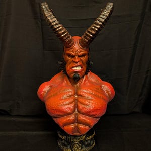 May include: A red-skinned Hellboy bust featuring large, dark, curved horns, a muscular chest, and a goatee. The character has a stern facial expression with visible teeth. The base is black with gold-coloured lettering and a circular emblem design.