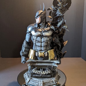 May include: A detailed Batman bust statue, featuring a black and gray color scheme with gold accents. The statue has a muscular physique and is mounted on a base with the text "Batman Arkham Knight". The bust is surrounded by bat-shaped wings.