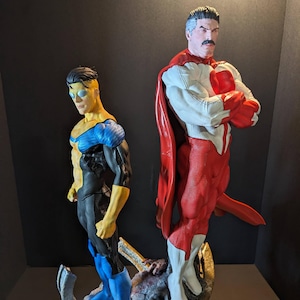 Op de afbeelding: Twee superheldenbeeldjes. Eén is geel, blauw en zwart, met een zonnebril. De andere is rood, wit en blauw, met een rode cape. Beide staan op een gedetailleerde, rotsachtige basis. De figuren staan in een heroïsche houding.