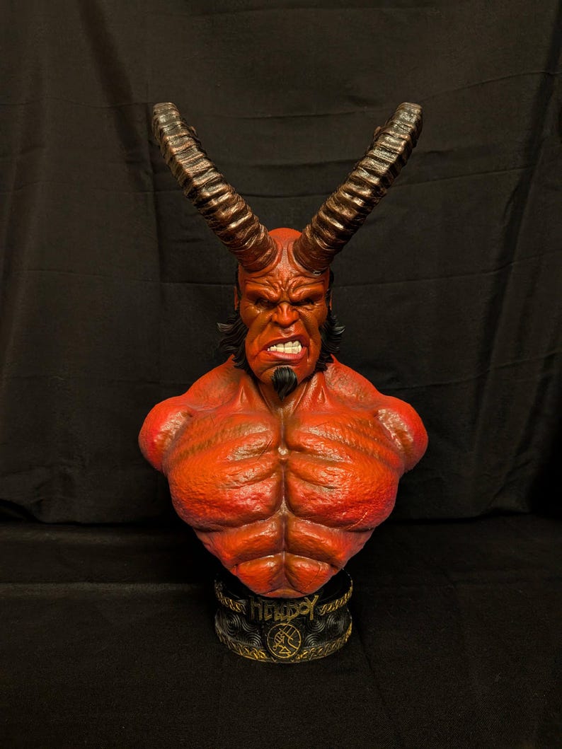 May include: A red-skinned Hellboy bust featuring large, dark, curved horns, a muscular chest, and a goatee. The character has a stern facial expression with visible teeth. The base is black with gold-colored lettering and a circular emblem design.