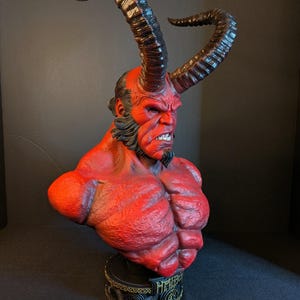 May include: A red-skinned Hellboy bust featuring large, curved, textured horns, a muscular torso, and a black goatee. The character has a stern facial expression with visible teeth. The base is black with gold-colored trim and the text HELLBOY.