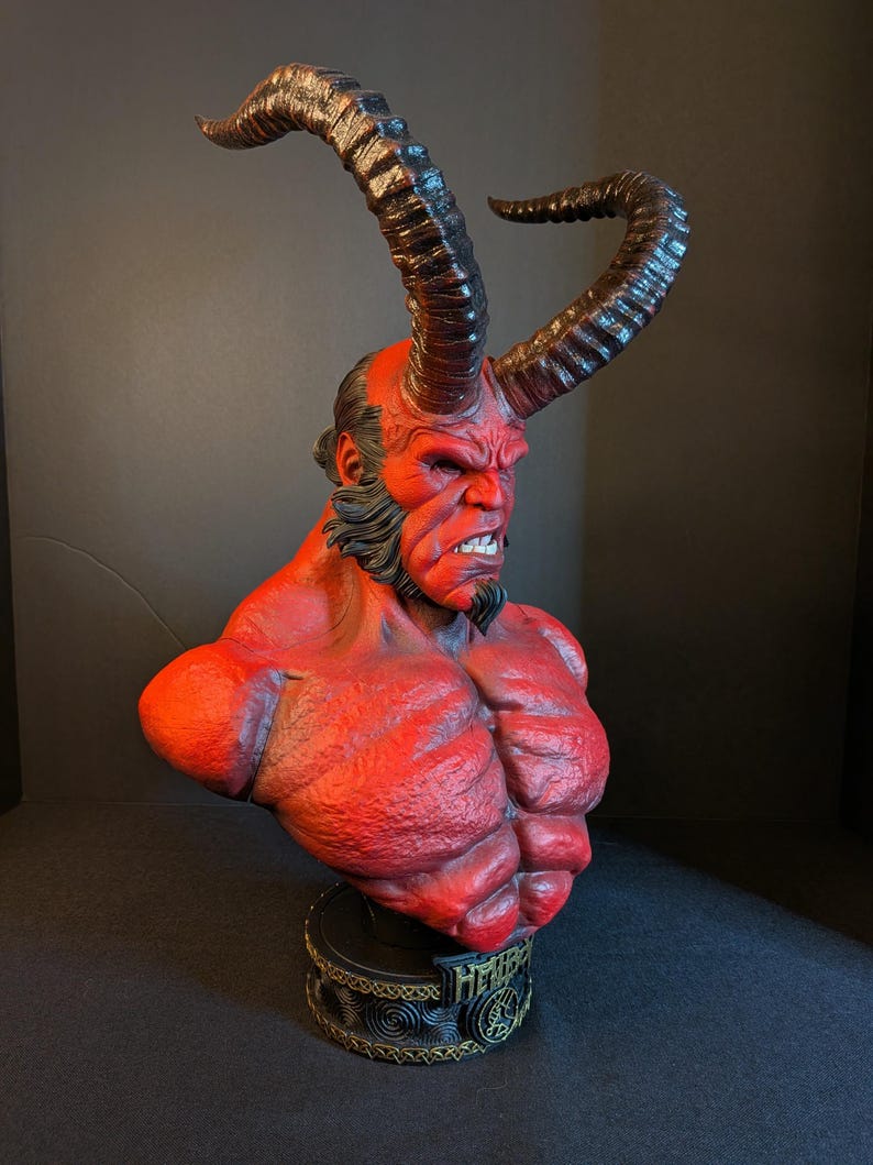 May include: A red-skinned Hellboy bust featuring large, curved, textured horns, a muscular torso, and a black goatee. The character has a stern facial expression with visible teeth. The base is black with gold-colored trim and the text HELLBOY.