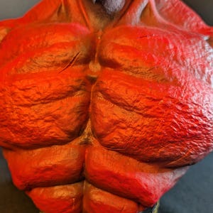 May include: A red-skinned, muscular torso figurine featuring a textured, rocky surface. The character has dark hair at the neck and sits atop a black base with gold-colored lettering that reads Hellboy, showcasing detailed anatomical definition.