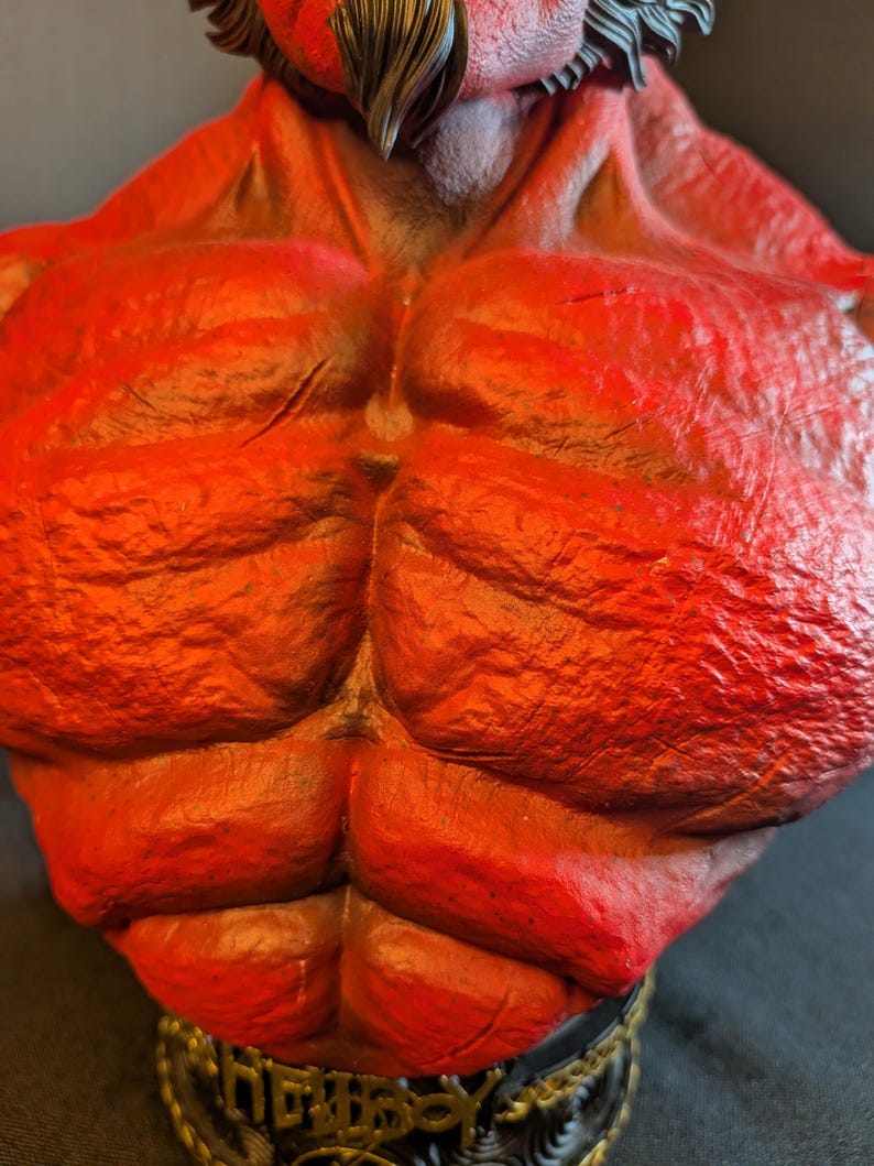 May include: A red-skinned, muscular torso figurine featuring a textured, rocky surface. The character has dark hair at the neck and sits atop a black base with gold-colored lettering that reads Hellboy, showcasing detailed anatomical definition.
