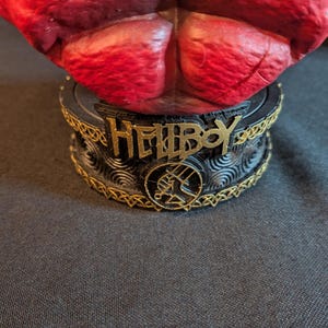 May include: A red textured figurine torso mounted on a circular black base. The base features the gold-colored text HELLBOY, a circular emblem with a hand symbol, and intricate gold-colored Celtic-style knotwork trim around the top and bottom edges.