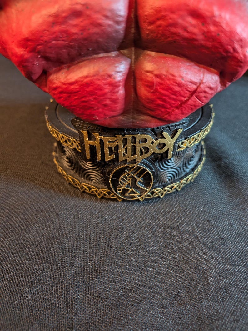 May include: A red textured figurine torso mounted on a circular black base. The base features the gold-colored text HELLBOY, a circular emblem with a hand symbol, and intricate gold-colored Celtic-style knotwork trim around the top and bottom edges.