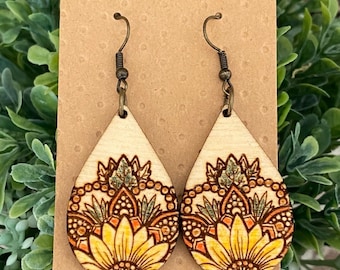 May include: A pair of wooden teardrop-shaped earrings with a floral design. The earrings are painted with yellow, orange, green, and brown colors. The earrings are hanging from a brown card.