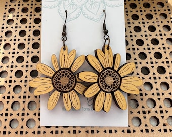 May include: A pair of wooden sunflower earrings with brown stems and gold-colored petals. The earrings are on a white card with a blue mandala design.
