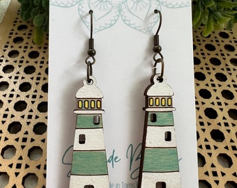 May include: A pair of wooden lighthouse earrings with white and green stripes. The earrings have a gold hook and are on a white card with a mandala design.