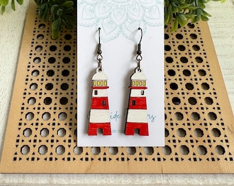 May include: A pair of red and white lighthouse earrings with yellow windows. The earrings are made of wood and have a brown metal hook.
