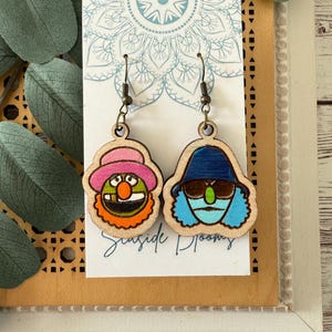 May include: Wooden earrings featuring two cartoon character designs. One earring depicts a character with a pink hat and orange mouth, the other with a blue hat and sunglasses. The earrings are on a white card with the text "Seaside Blooms".