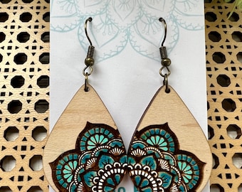 May include: Teal and brown mandala wood earrings with a teardrop shape and gold hooks. The earrings are displayed on a white card with the text "Lightweight wood earrings".