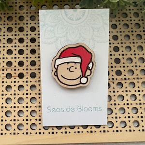 May include: A wooden pin with a cartoon character wearing a red Santa hat. The pin is on a white card with the text "Seaside Blooms".