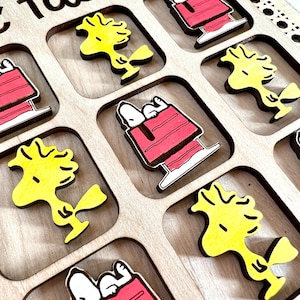 May include: A wooden tic-tac-toe game with yellow and red cartoon characters. The game board has nine squares, and each square has a different character. The characters are all from the Peanuts comic strip.