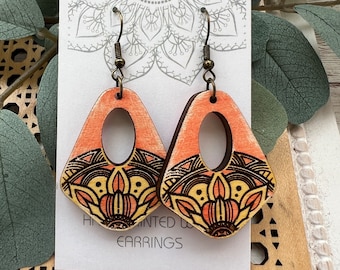 May include: A pair of hand-painted wooden earrings with a peach and yellow mandala design. The earrings are displayed on a white card with the text "Hand Painted Wood Earrings".