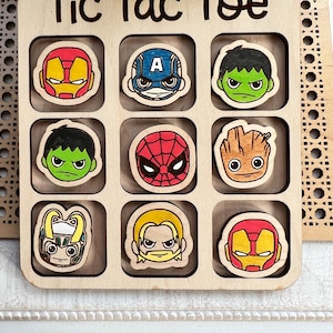 May include: Wooden tic-tac-toe game with superhero character illustrations. The game board has nine squares with images of Iron Man, Captain America, Hulk, Spiderman, Groot, Loki, Thor, and Iron Man. The game board is labeled "Tic Tac Toe".