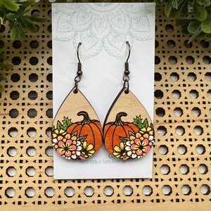 May include: Wooden teardrop earrings featuring a pumpkin design with orange, yellow, and pink flowers. The earrings have bronze-colored hooks and are set against a light-colored background. The text "Handmade in Toms River, NJ" is visible.