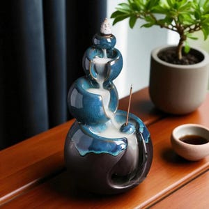 Puede incluir: Fuente de cascada de incienso de cerámica azul. El diseño es en cascada con un acabado brillante y una base azul oscuro. Se coloca una varilla de incienso en la fuente y el humo fluye por los niveles. Una pequeña planta y una taza están en el fondo.