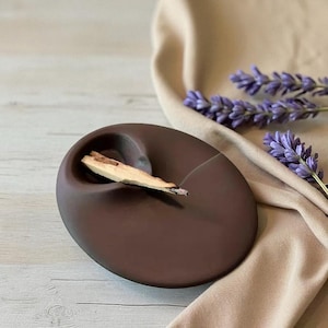 Könnte beinhalten: Eine dunkelbraune, ovale Keramikschale mit einer geschwungenen Vertiefung, die ein Stück helles Holz enthält. Die Schale steht auf einer hellen Oberfläche, mit einem beigen Stoff und Zweigen von lila Lavendel im Hintergrund.