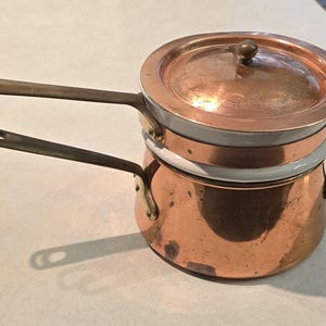 May include: A double boiler set featuring a copper exterior with a white enamel interior lining. The set includes a bottom pot with a side handle and a smaller top pot with a matching copper lid and a long handle extending from the side.