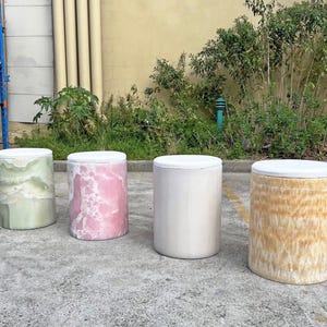 May include: Four cylindrical stools with white tops. The stools are in various colours and textures: light green, pink marble, light grey, and golden brown. They are made of stone or similar material and are suitable for indoor or outdoor use.
