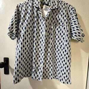 May include: A white short-sleeved button-down shirt with a blue and white geometric pattern. The shirt has a collar and a pocket on the left side. The shirt is hanging on a wooden hanger.