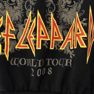 May include: Black sleeveless shirt featuring the band name "Def Leppard" in yellow and red, with a decorative design above. The text "World Tour 2008" is printed below the band name in gray.