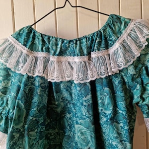 Könnte beinhalten: Eine tealblaue Bluse mit Blumendruck und weißer Spitzenborte am Ausschnitt und an den Ärmeln.