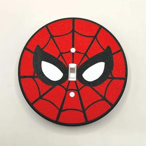 May include: A round, red light switch cover featuring a black spiderweb design and black eye shapes. The switch cover has a white toggle switch and is mounted on a white wall.