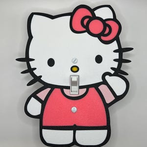 以下が含まれることがあります： 漫画のキャラクターデザインが施された装飾的なライトスイッチカバー。カバーは白で、ピンクのリボン、ピンクのドレス、黒いアウトラインが付いています。ライトスイッチは標準的なトグルスタイルです。