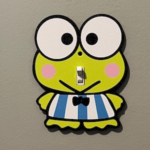 Puede incluir: Una cubierta decorativa para interruptor de luz con forma de rana de dibujos animados. La rana tiene grandes ojos blancos, mejillas rosadas y una camisa a rayas azules y blancas. El interruptor está en el centro de la cara de la rana.