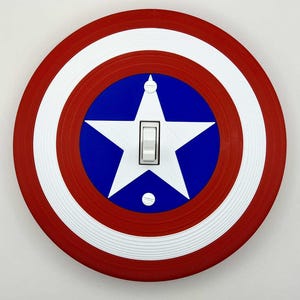 Puede incluir: Una cubierta de interruptor de luz redonda diseñada para parecerse al escudo del Capitán América. La cubierta presenta un anillo exterior rojo, un anillo interior blanco y un centro azul con una estrella blanca. El interruptor es de tipo palanca estándar.