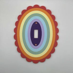 May include: A decorative light switch cover featuring a rainbow design. The cover has an oval shape with a scalloped red border. Inside, concentric ovals display the colors of the rainbow, with a purple center for the switch opening. Two small holes are visible.