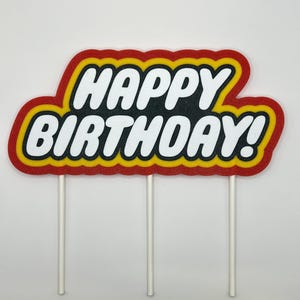 May include: A colorful cake topper with the words "HAPPY BIRTHDAY!" in white, outlined in yellow and red. The topper is mounted on three white sticks, perfect for decorating a birthday cake.
