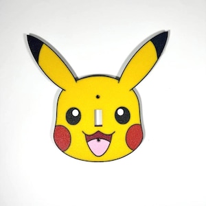 Op de afbeelding: Een gele Pikachu-vormige lichtschakelaar cover met zwartgepunte oren, zwarte ogen, rode wangen en een roze tong. De cover heeft een rechthoekige opening voor de lichtschakelaar, tegen een witte achtergrond.