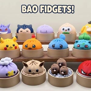 May include: A collection of fourteen colourful character-themed bao fidget toys, each resting in a small circular tan container. The figures feature various facial expressions and colours, including yellow, purple, blue, and red. The text BAO FIDGETS! is visible.