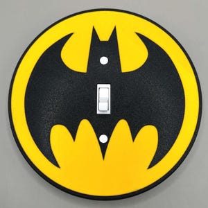 May include: A circular light switch cover featuring a black bat silhouette centred on a bright yellow background. The cover has a textured finish, a central rectangular cutout for a toggle switch, and two small circular holes for mounting screws.