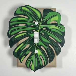 Peut inclure: Un cache-interrupteur décoratif avec un motif de feuille de monstera verte. La feuille présente différentes nuances de vert et se trouve sur une plaque d'interrupteur beige. L'interrupteur et les vis sont blancs.