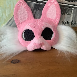 May include: A pink felt bunny mask with large black mesh eye openings and white faux fur trim. The mask has decorative details on the ears and nose. The artist's signature, "Kait Rob", is visible on the right side.