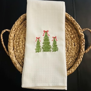 Puede incluir: Una toalla de cocina blanca con tejido de gofre con tres árboles de Navidad verdes bordados, cada uno coronado con un lazo rojo. La toalla está doblada y descansa en una cesta redonda tejida con asas.