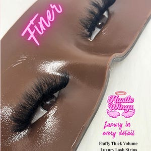以下が含まれることがあります： 茶色の表面のクローズアップで、太くてふわふわした黒いつけまつげがペアになっています。「Finer」という言葉はピンク色のスクリプトで書かれています。「Hustle Wings」というテキストは「Luxury in every detail」というテキストの上にあります。「Fluffy Thick Volume Luxury Lash Strips 長さ：25mm」というテキストも表示されています。